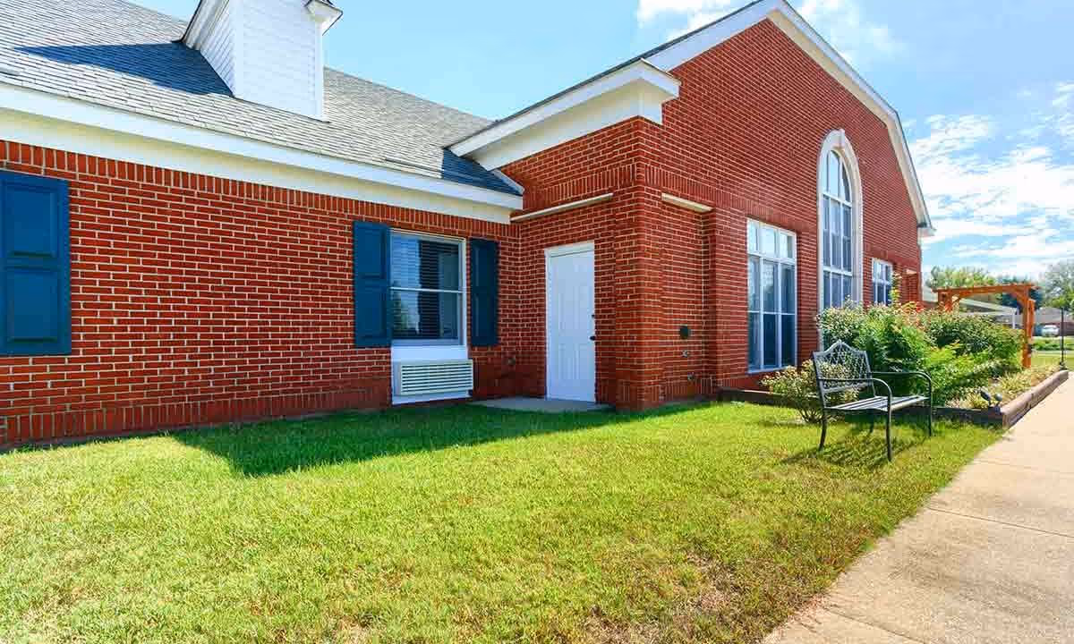 Exterior view of a red brick building with blue shutters on the windows, a white door, and a well-maintained lawn with a black metal bench and some bushes under a partly cloudy sky.