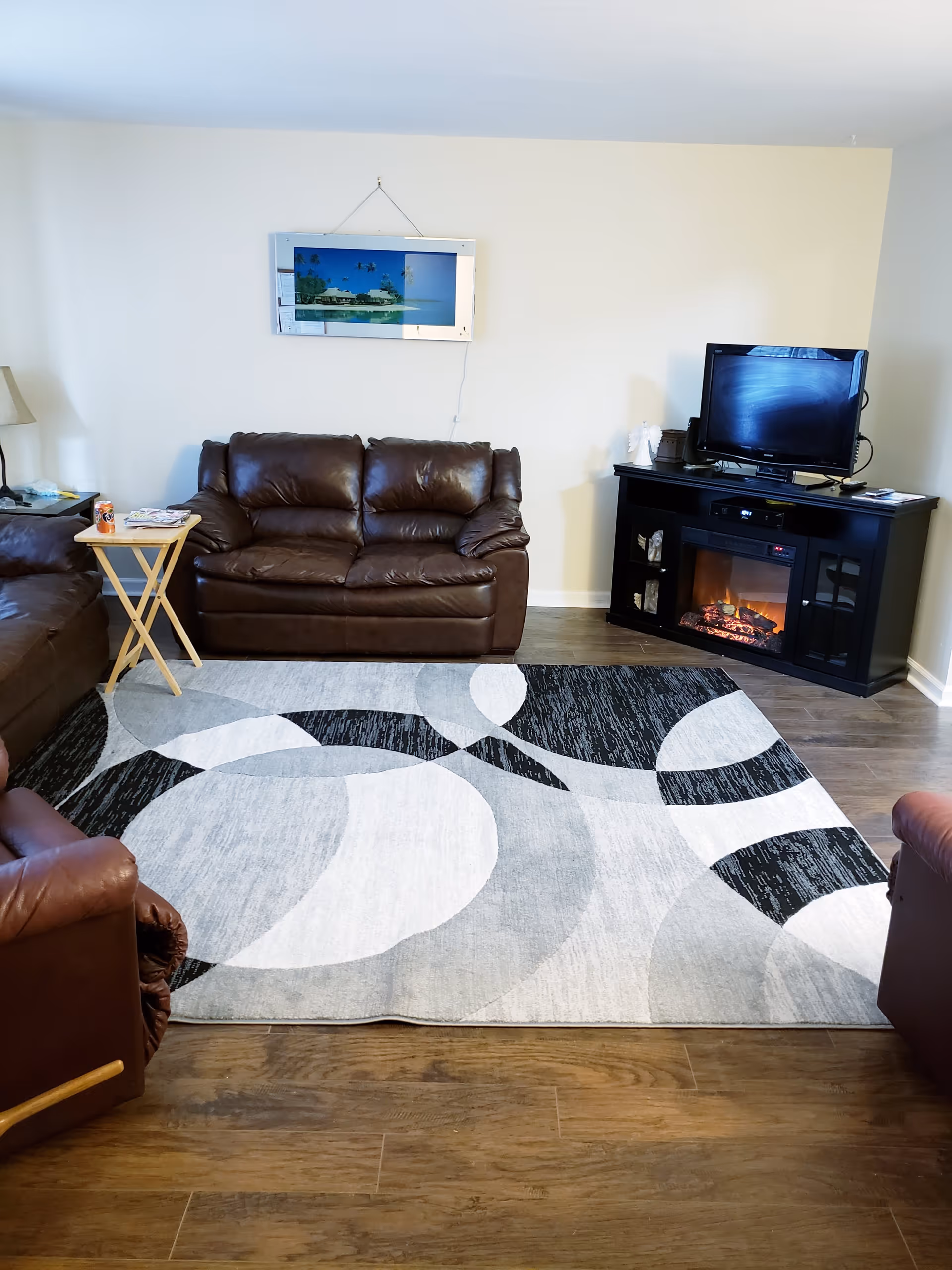 A living room with brown leather sofas arranged around a black and white patterned area rug. There is a small wooden side table with a can of soda and some magazines on it. On the right side, a black entertainment unit with an electric fireplace and a TV on top is visible. A framed picture of a tropical beach scene hangs on the wall above the sofa.