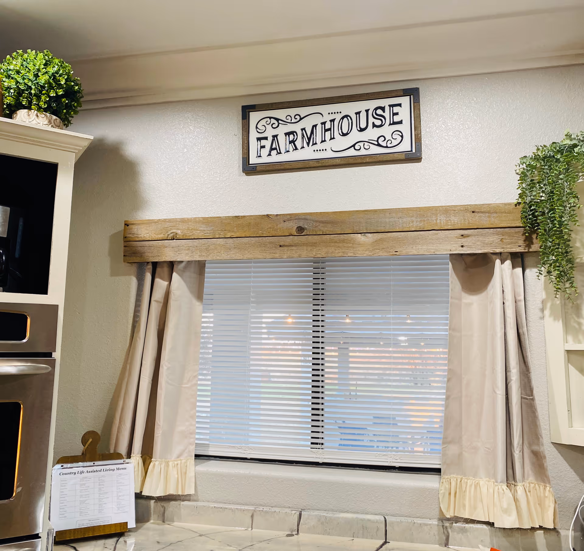 Interior view of a kitchen area with a window covered by beige curtains and white blinds. Above the window is a wooden valance and a decorative sign that reads 'FARMHOUSE'. On the left side, there is a built-in oven and a small menu stand labeled 'Country Life Assisted Living Menu'. There are green potted plants on top of the cabinets on both sides of the window.
