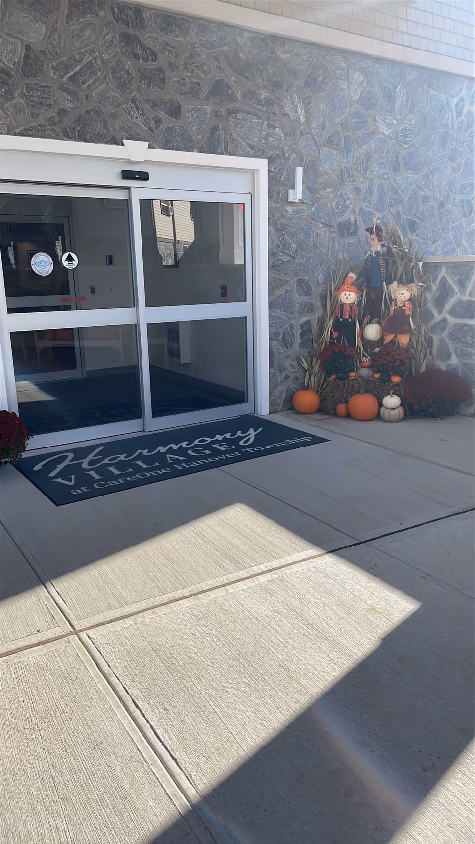 Entrance to Harmony Village at CareOne Hanover Township with automatic sliding glass doors, a stone wall exterior, a welcome mat with the facility name, and fall-themed decorations including pumpkins, scarecrows, and dried corn stalks.
