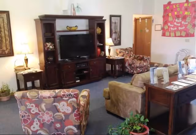 A cozy living room with floral patterned armchairs, a beige armchair, a wooden entertainment center with a flat-screen TV, side tables with lamps, framed artwork on the walls, and a bulletin board with colorful decorations. There is also a wooden table with brochures and a potted plant in the foreground.
