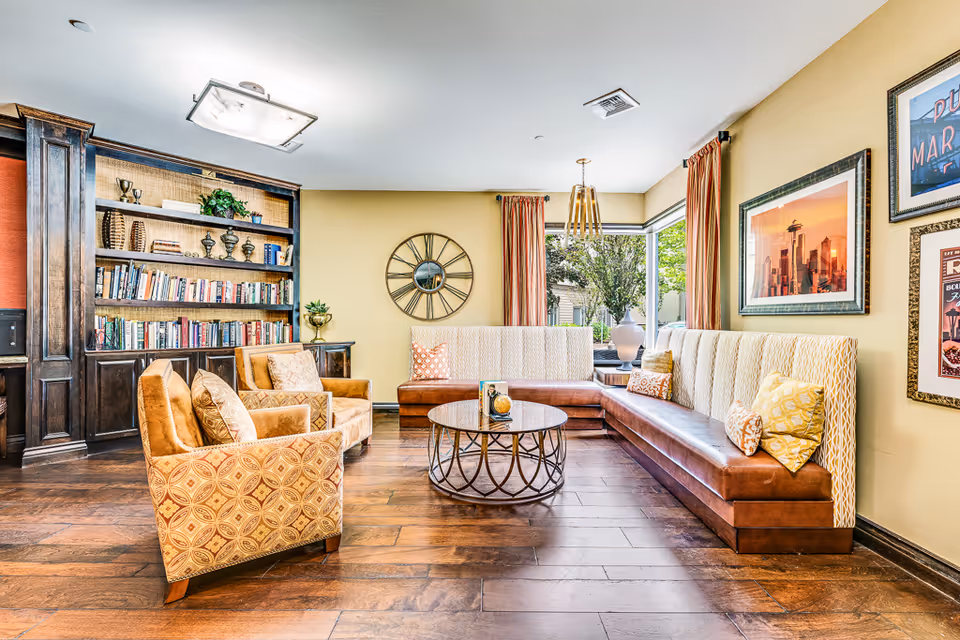 A bright and cozy living room area with wooden flooring, two patterned armchairs, a round coffee table, and a long cushioned bench with decorative pillows. There is a large built-in bookshelf filled with books and decorative items, a wall clock, framed pictures on the walls, and large windows with curtains letting in natural light.