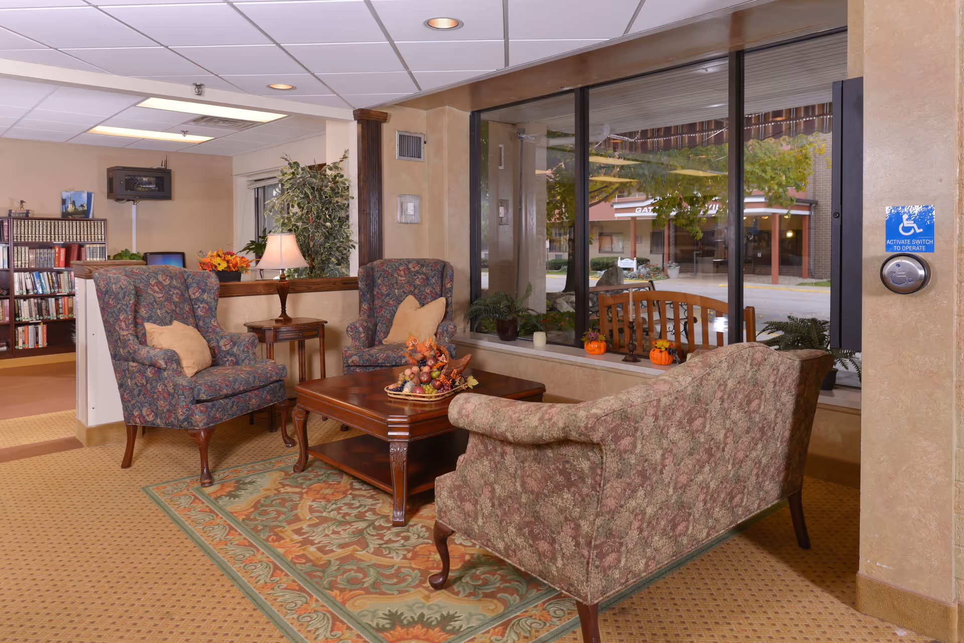 A cozy senior living facility common area with floral upholstered armchairs and a matching sofa arranged around a wooden coffee table with a decorative centerpiece. A patterned rug lies beneath the furniture. Large windows reveal an outdoor view with trees and a bench. A bookshelf and a lamp on a side table are visible in the background.