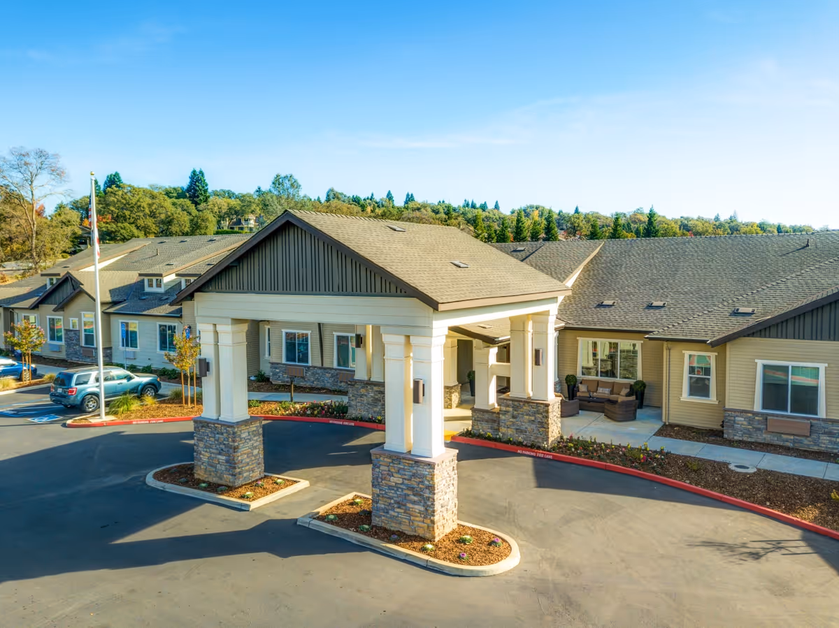 Exterior view of The Pavilion at El Dorado Hills senior living facility showing the main entrance with a covered drop-off area supported by stone and white pillars. The building has beige siding with stone accents and multiple windows. There are a few parked cars and landscaped areas with small trees and shrubs. The sky is clear and blue.
