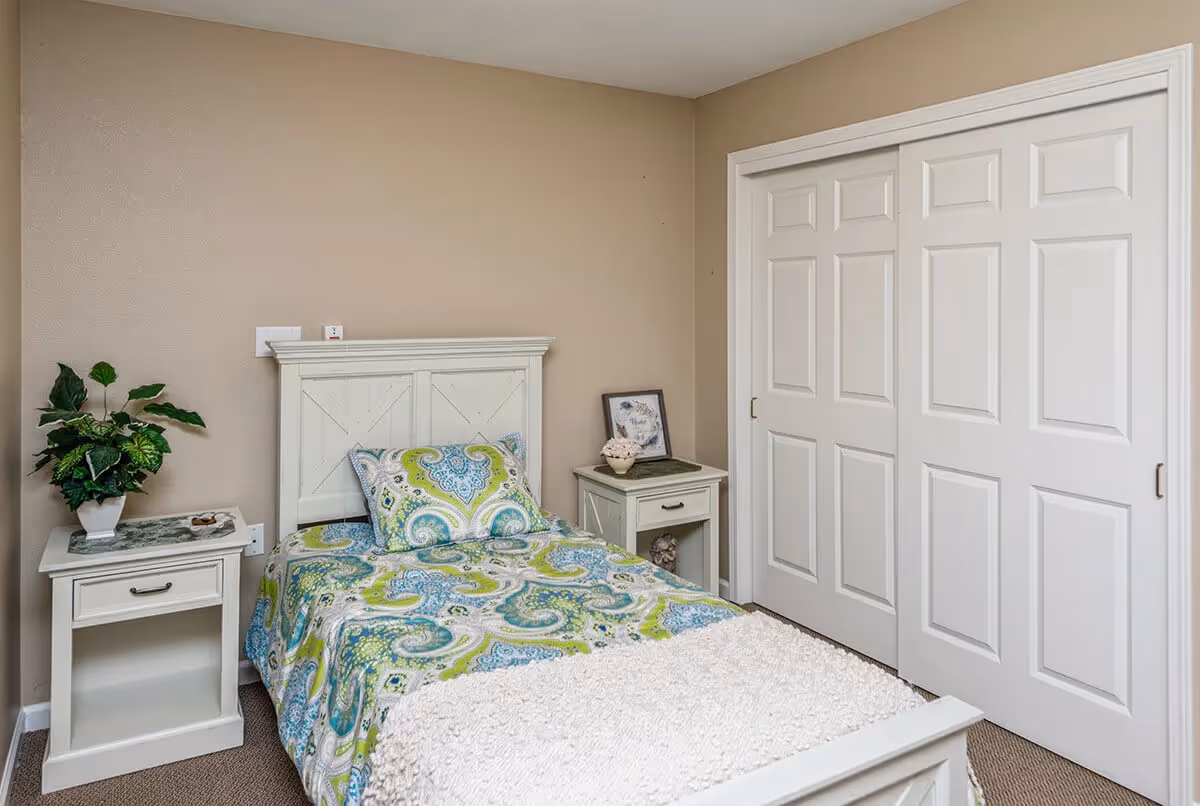 A small bedroom with a single bed featuring a colorful paisley-patterned bedspread in shades of green, blue, and white. The bed has a white wooden headboard and a matching white nightstand on each side. One nightstand holds a potted plant and a small dish, while the other has a framed picture and a decorative item. There is a large white closet with sliding doors on the right side of the room. The walls are beige and the floor is carpeted.