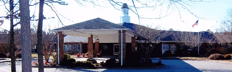 Front exterior view of a single-story brick building with a covered entrance supported by brick columns. There are leafless trees and landscaped bushes around the building, and an American flag is visible on the right side.
