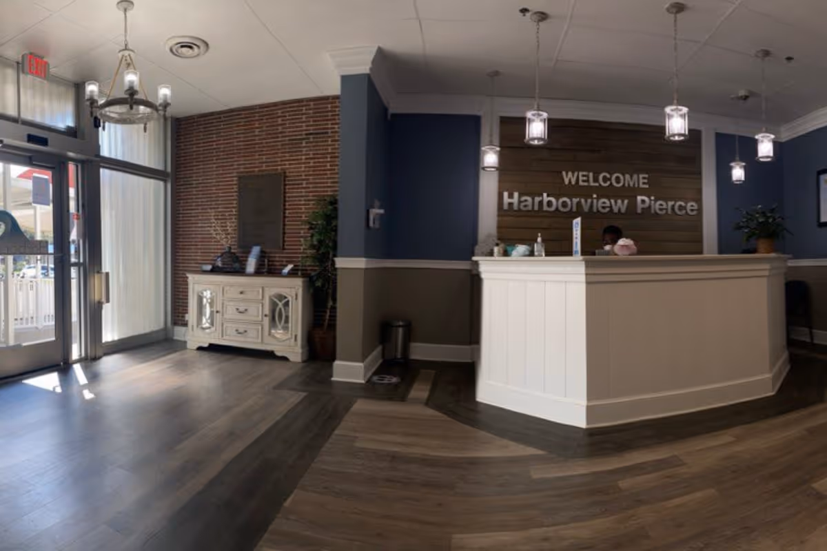 Reception area of Harborview Health Systems Pierce County featuring a white reception desk with a person seated behind it, a wooden welcome sign on the wall that reads 'WELCOME Harborview Pierce', pendant lights hanging from the ceiling, a small white cabinet with decorative items, and a glass entrance door letting in natural light.