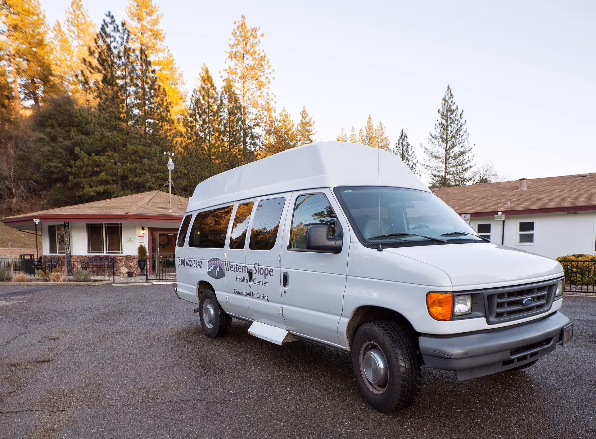 A white passenger van with the logo and name 'Western Slope Health Center' parked outside a single-story building surrounded by trees during daylight.