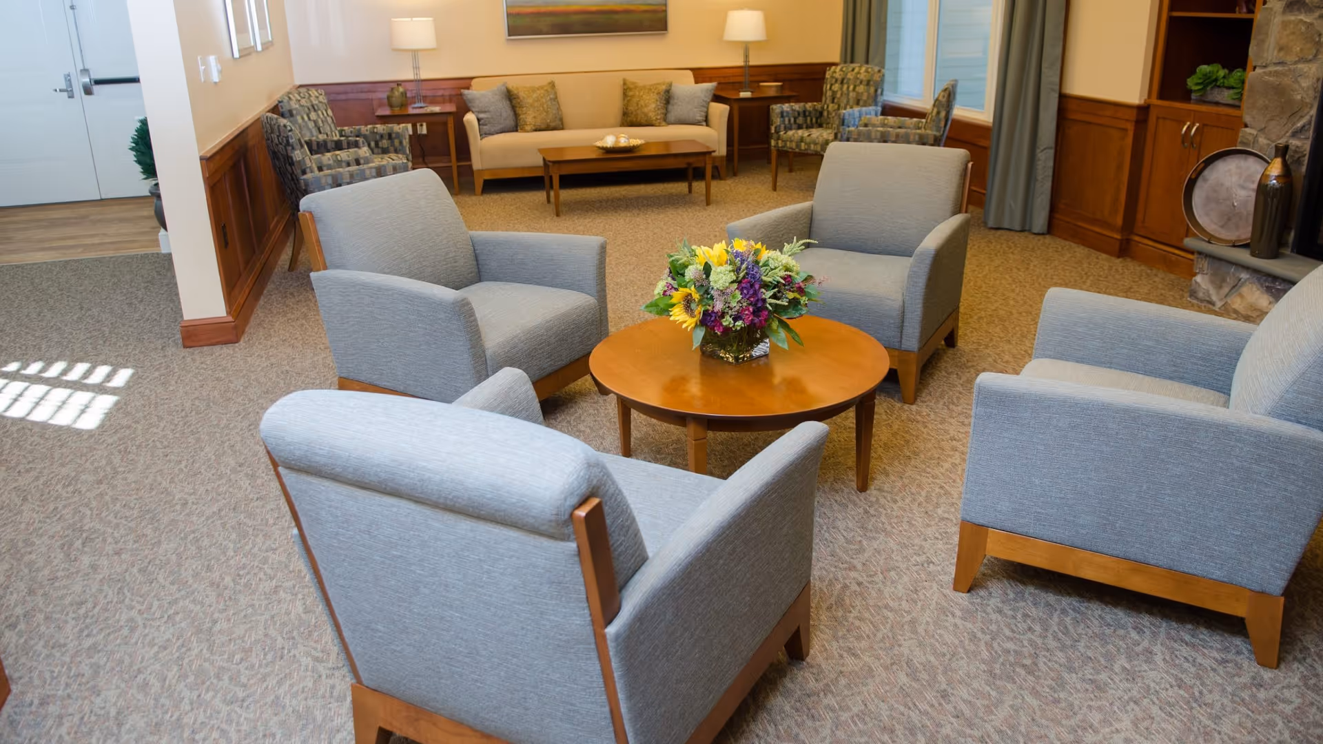 A cozy living room area with four gray armchairs arranged around a round wooden coffee table with a colorful flower arrangement. In the background, there is a beige sofa with cushions, two patterned armchairs, wooden side tables with lamps, a stone fireplace, and built-in wooden shelves with decorative items.