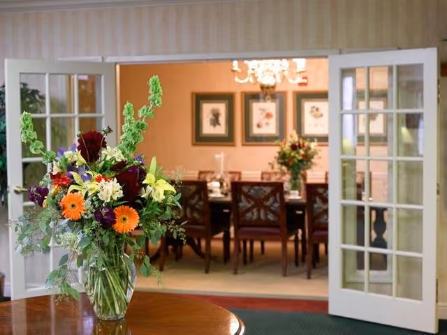 A large glass vase filled with a colorful bouquet of flowers sits on a wooden table in the foreground. Behind it, open white French doors lead into a dining room with a long table surrounded by chairs. The dining room walls are decorated with framed artwork and a chandelier hangs from the ceiling.