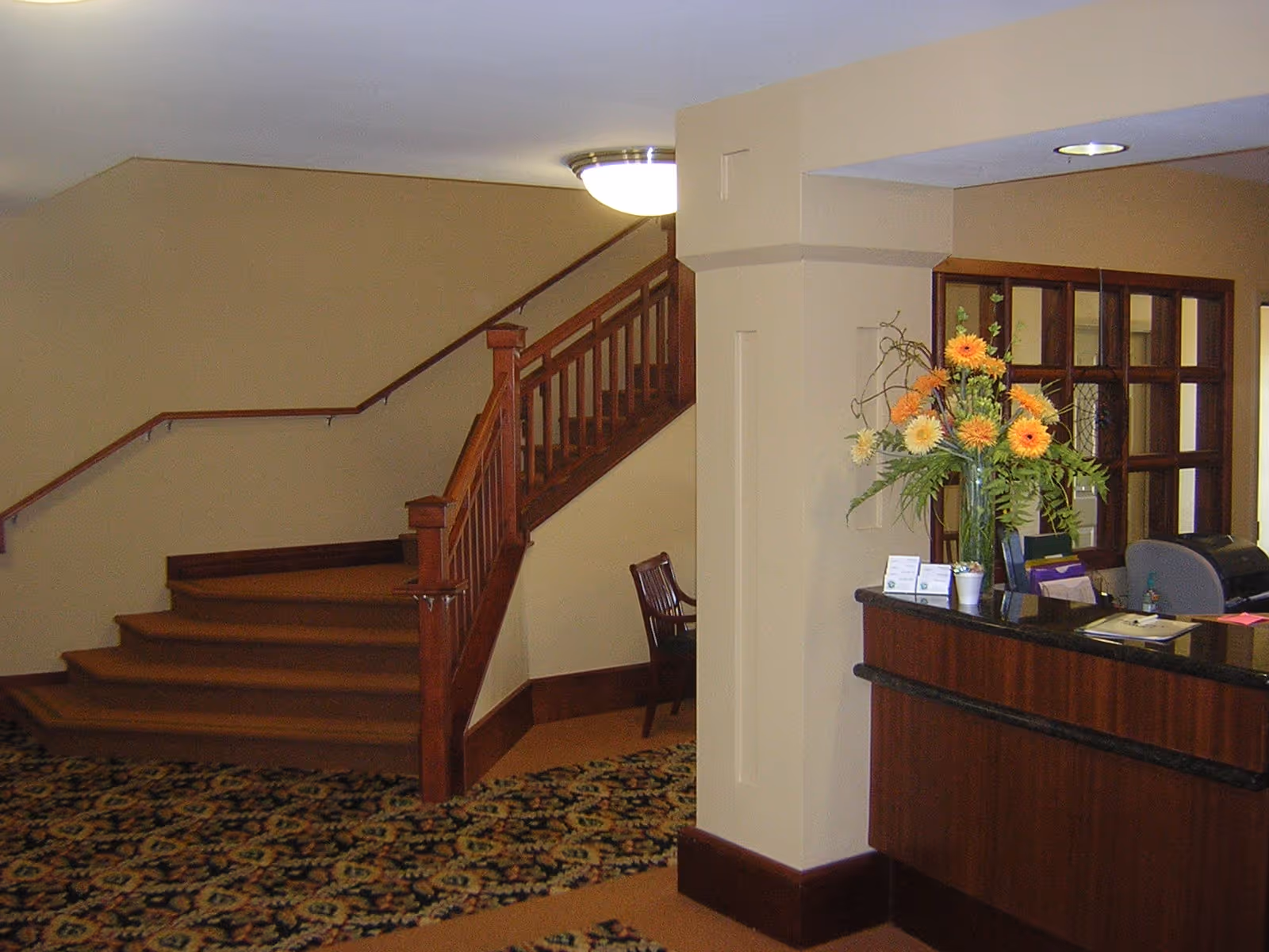Interior view of a senior living facility lobby area with a wooden staircase, a reception desk with a vase of flowers, and a chair in the background. The floor is covered with patterned carpet and the walls are painted beige.
