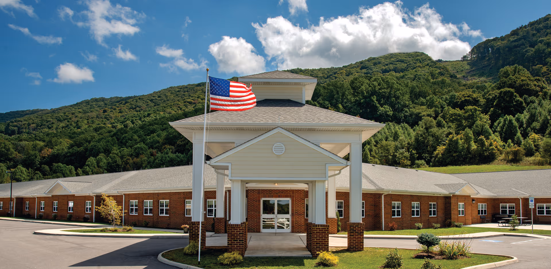 Front exterior view of Mercer Healthcare Center, a single-story brick building with a covered entrance supported by white columns. An American flag is flying on a flagpole in front of the entrance. The building is set against a backdrop of green, forested hills under a blue sky with scattered clouds.