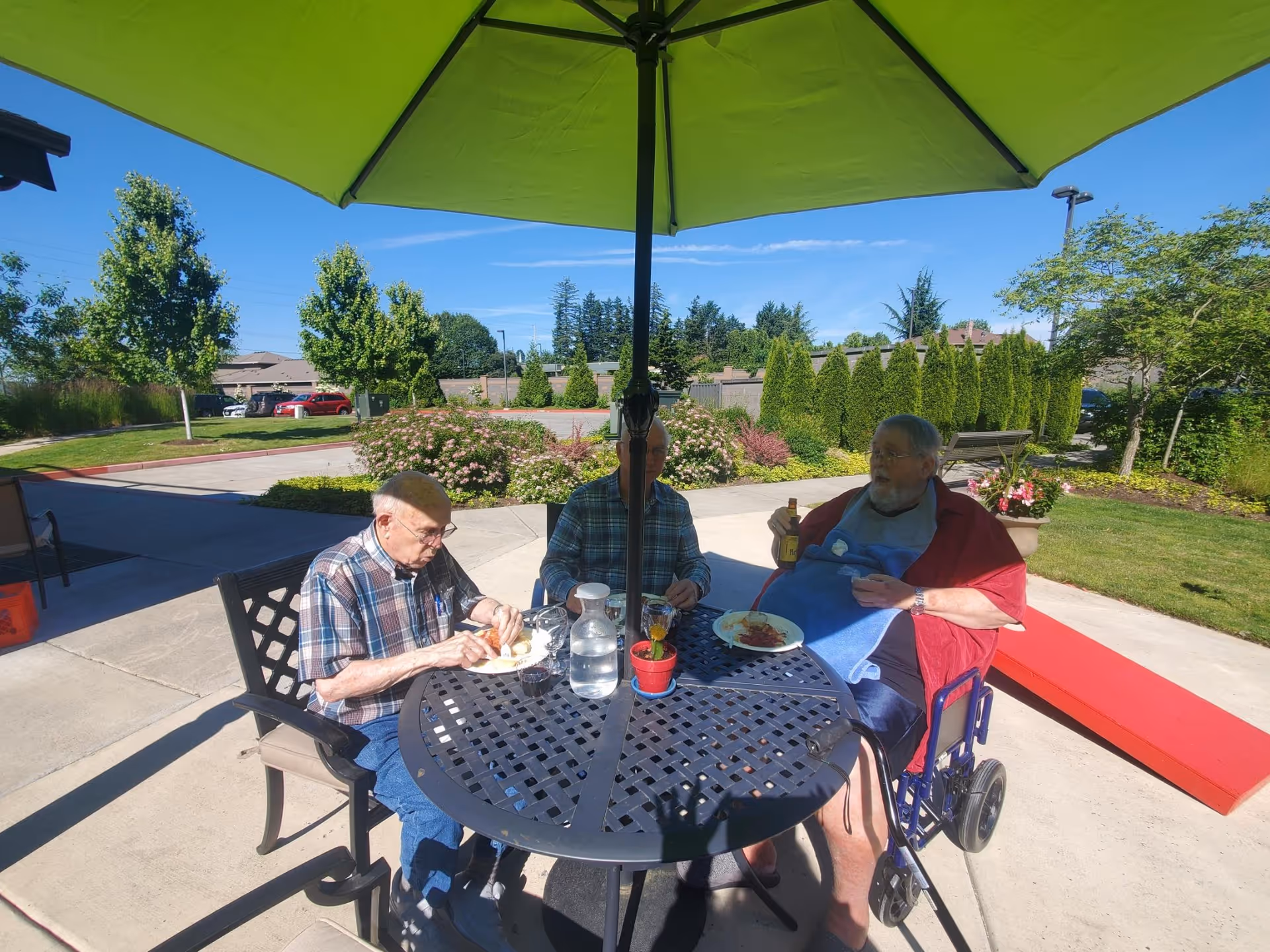 Three elderly men sitting around a round outdoor table under a large green umbrella. They are eating a meal together in a garden area with trees, bushes, and a clear blue sky in the background. One man is in a wheelchair with a blue bib and holding a bottle, while the other two men are seated on chairs.