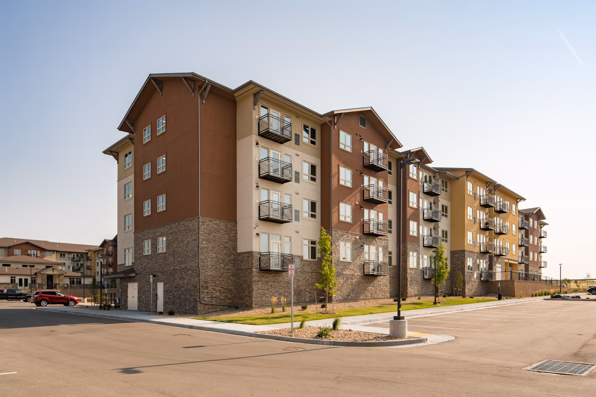 Exterior view of a multi-story residential building with balconies, stone and stucco facade, and a parking lot in front under a clear sky.