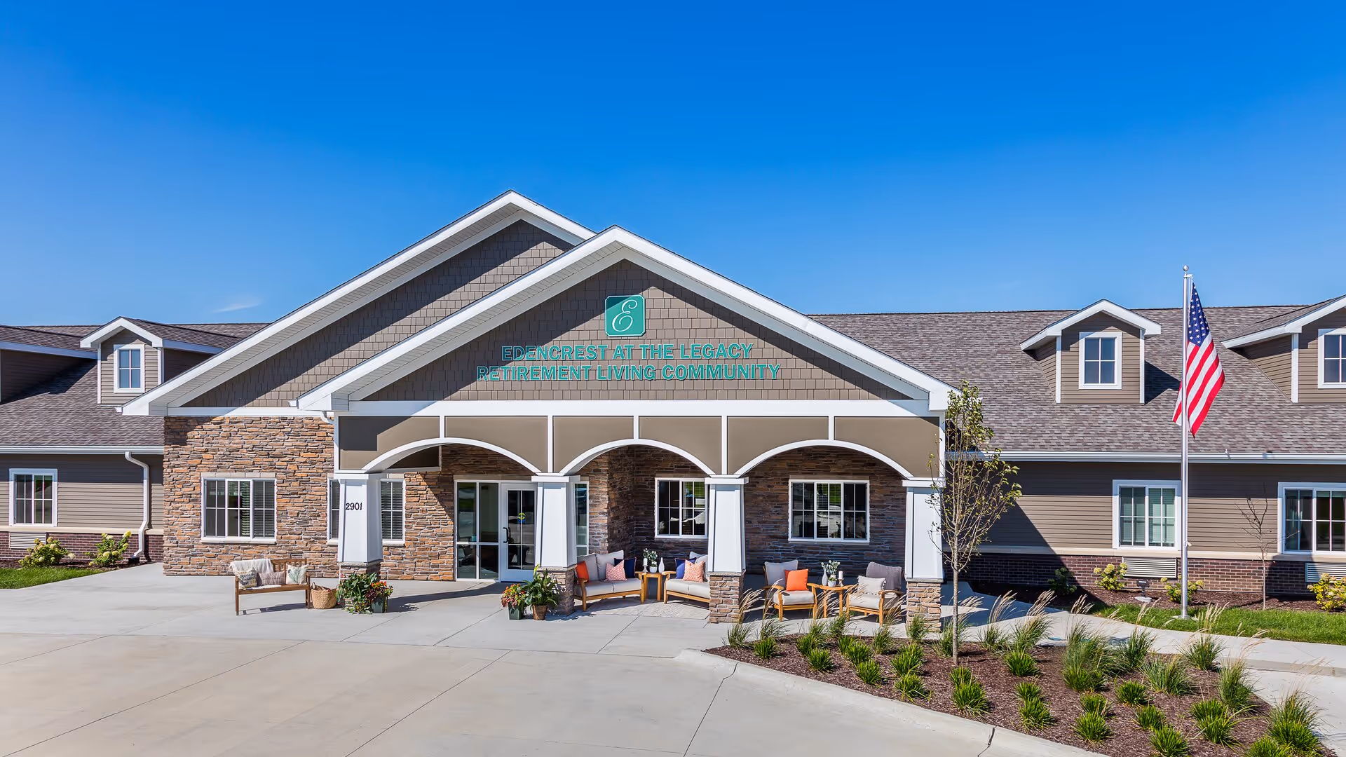 Front exterior view of Edencrest at The Legacy retirement living community building with a covered entrance, outdoor seating area with chairs and tables, an American flag on a flagpole, and clear blue sky.
