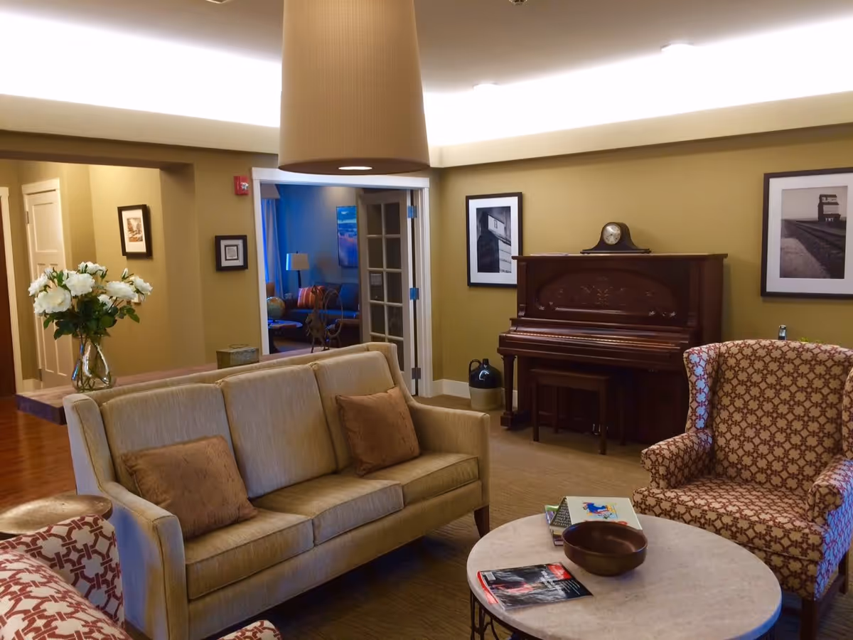 A cozy living room in an assisted living facility featuring a beige sofa with two brown pillows, a patterned armchair, a round coffee table with books and a bowl, a wooden upright piano with a clock on top, framed black and white photographs on the wall, and a vase with white flowers on a side table. The room has warm lighting and an open doorway leading to another room with blue walls and additional seating.