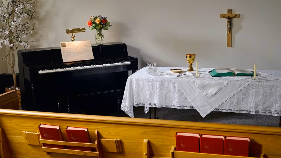 Interior of a small chapel or prayer room with wooden pews holding red hymnals, a black piano with sheet music and a vase of flowers on top, and a table covered with a white lace cloth displaying religious items including candles, a chalice, a bowl, and an open book. A crucifix is mounted on the wall above the table.
