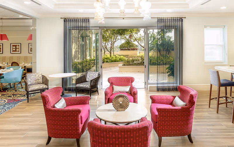 A bright and inviting common area with four red upholstered armchairs arranged around a round coffee table with a decorative centerpiece. Behind the seating area is a large sliding glass door with sheer curtains, leading to an outdoor garden space with greenery and trees. To the left, there is a dining area with tables, chairs, and blue and gray upholstered seating. To the right, there is a high table with bar stools near a window.
