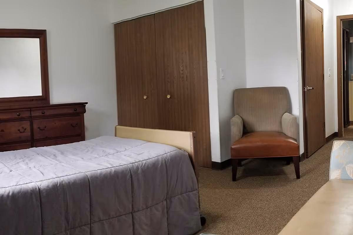 A bedroom in a senior living facility featuring a bed with a gray comforter, a wooden dresser with a mirror, a brown armchair, a closet with wooden sliding doors, and a doorway leading to another room.