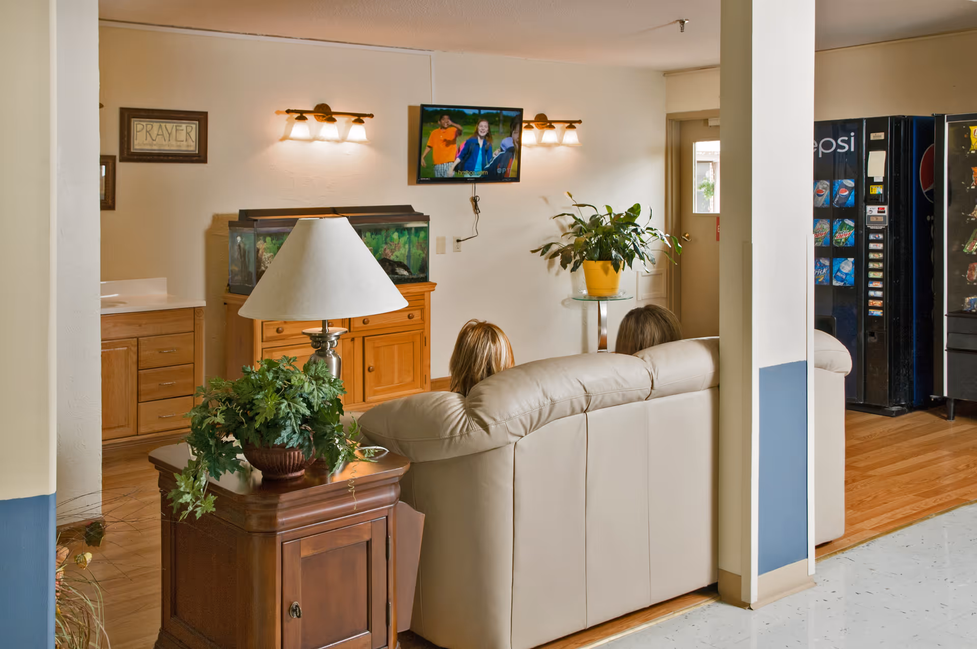 A beige couch in a care center lounge faces a wall-mounted TV with a fish tank, plants, and vending machines visible in the background.
