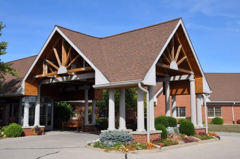 Covered entryway with white columns, exposed timber trusses and flowerbeds in front of a single-story brick senior living building.