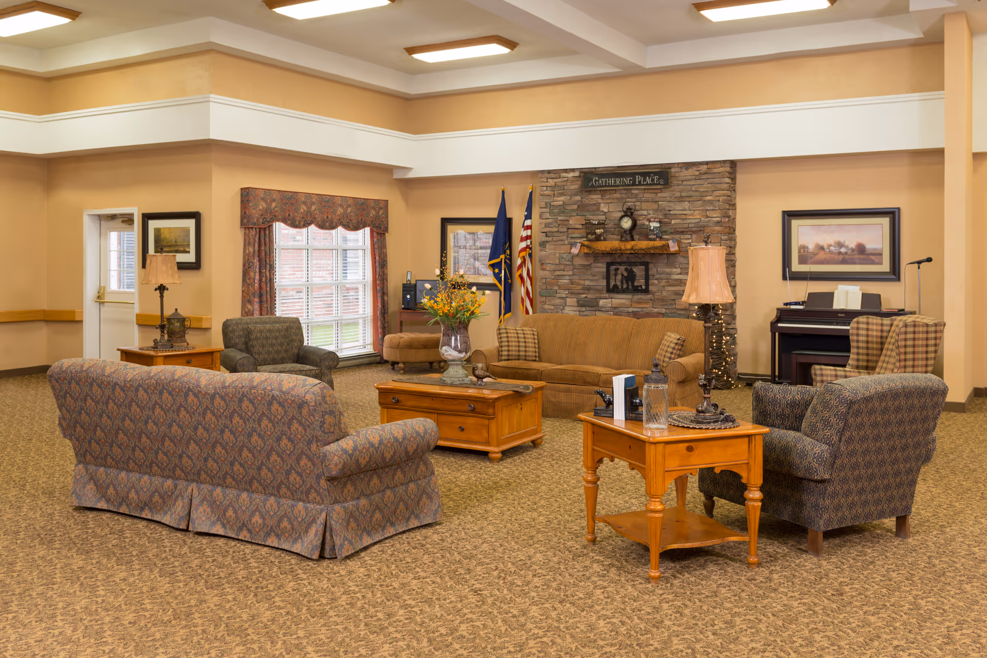 A cozy living room area in a senior living facility with patterned armchairs and sofas arranged around wooden coffee and side tables. The room features a stone fireplace with a sign above it that reads 'Gathering Place', two flags, framed artwork on the walls, a piano in the corner, and warm lighting from table lamps.