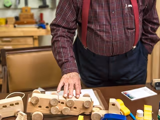 An elderly person wearing a checkered shirt and suspenders is standing behind a table with wooden toy vehicles and various small tools and objects on it.