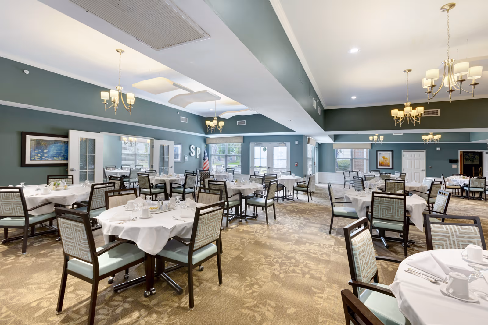 Spacious dining room with multiple round tables set with white tablecloths, wooden chairs, chandeliers, and teal walls.