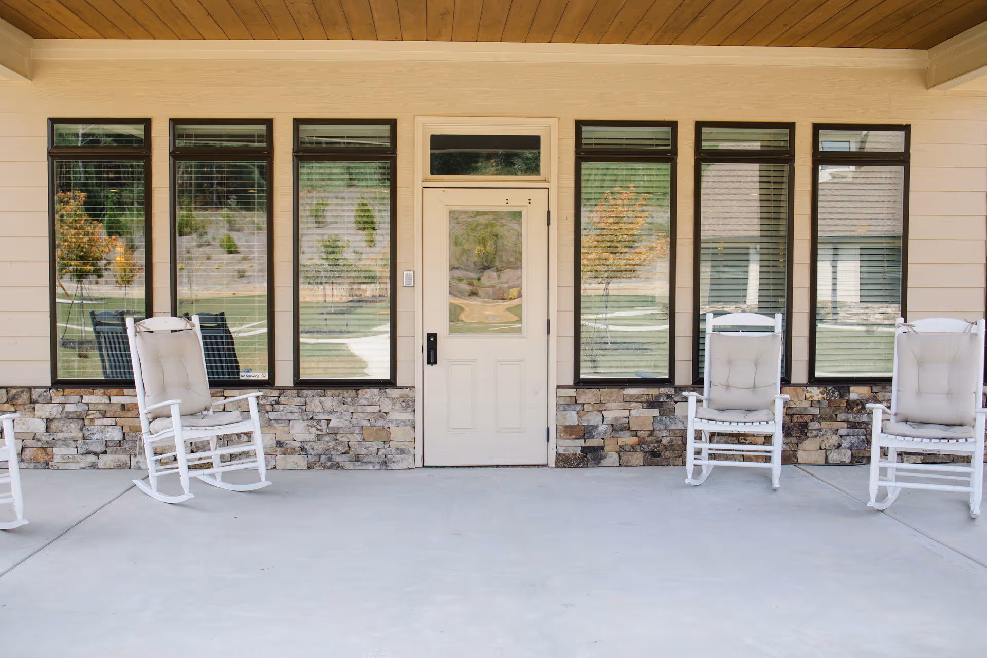 Covered porch area with four white cushioned rocking chairs placed in front of a beige wall with stone accents and five tall windows. A white door with a window at the top center is in the middle of the wall, reflecting the outdoor greenery and pathway.