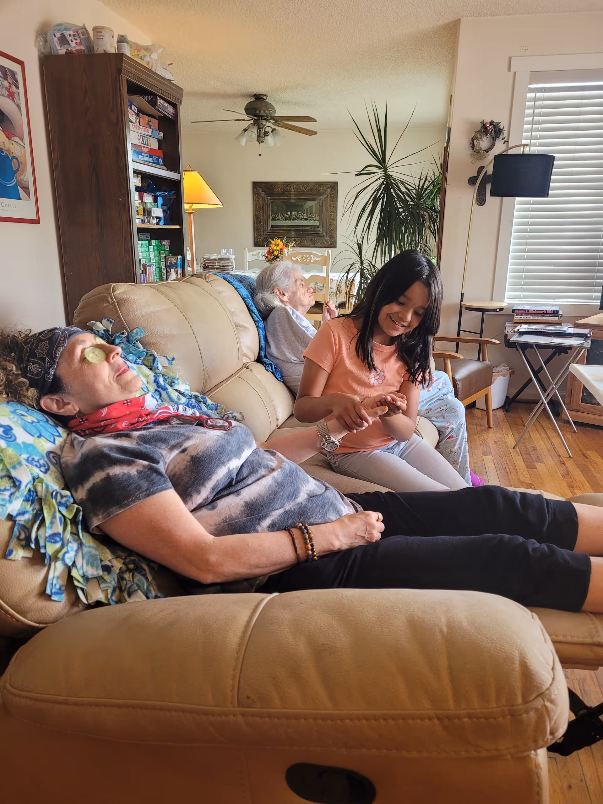 Three people in a cozy living room: a woman reclining with cucumber slices on her eyes, an elderly person seated on the couch, and a young girl painting nails.