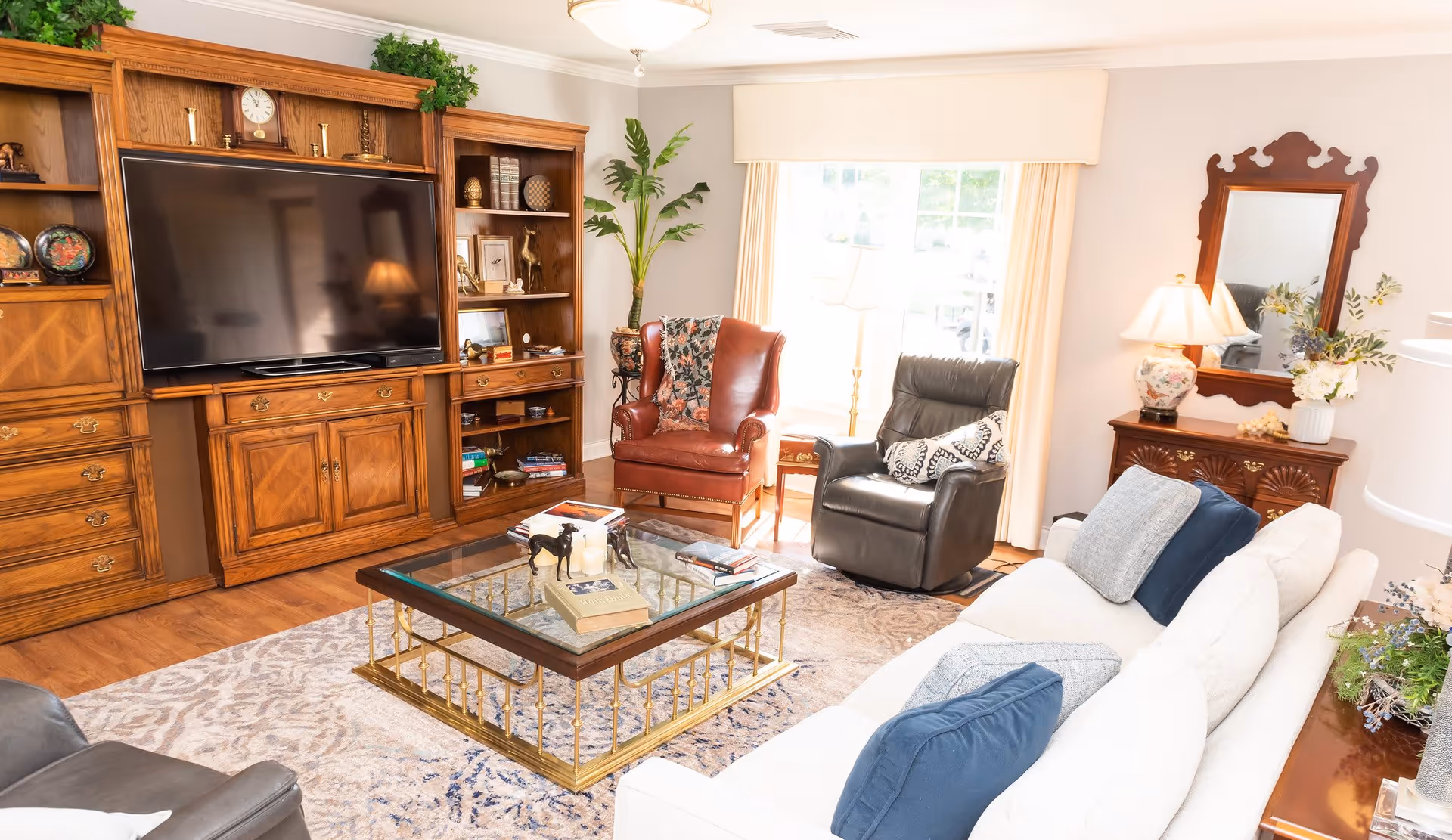 A bright and cozy living room with a large wooden entertainment center holding a flat-screen TV, a glass coffee table with books and decorative items, a white sofa with blue cushions, a brown leather armchair, a black recliner, a wooden side table with a lamp and flowers, and a large window with cream-colored curtains letting in natural light.