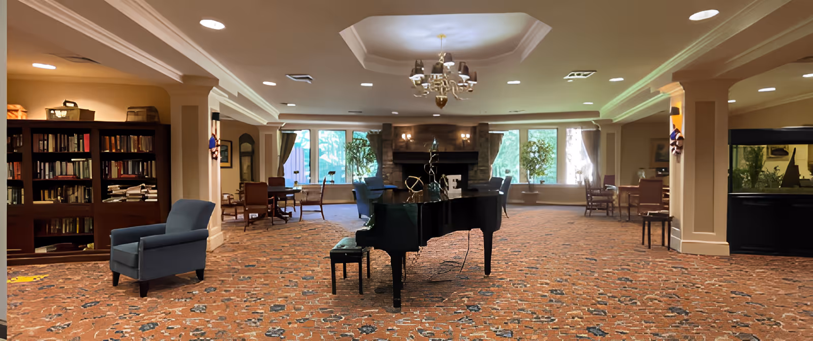 A spacious senior living facility common area with a grand piano in the center, surrounded by armchairs and tables. The room features large windows letting in natural light, a stone fireplace at the back, bookshelves on the left, and an aquarium on the right. The ceiling has recessed lighting and a chandelier.