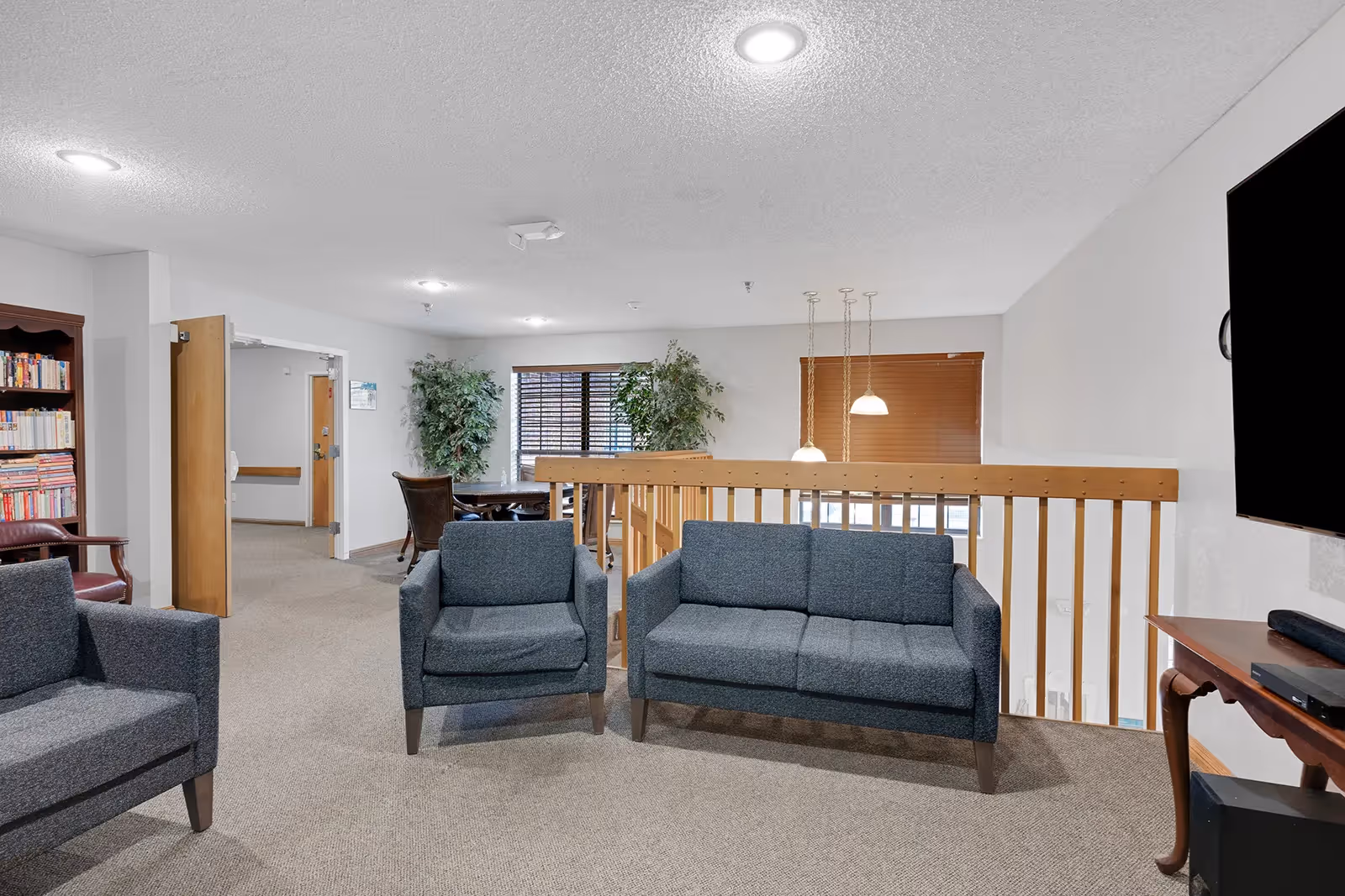 Communal seating area with gray sofas and armchairs overlooking a wooden railing, bookshelves, potted plants, and a wall-mounted TV.