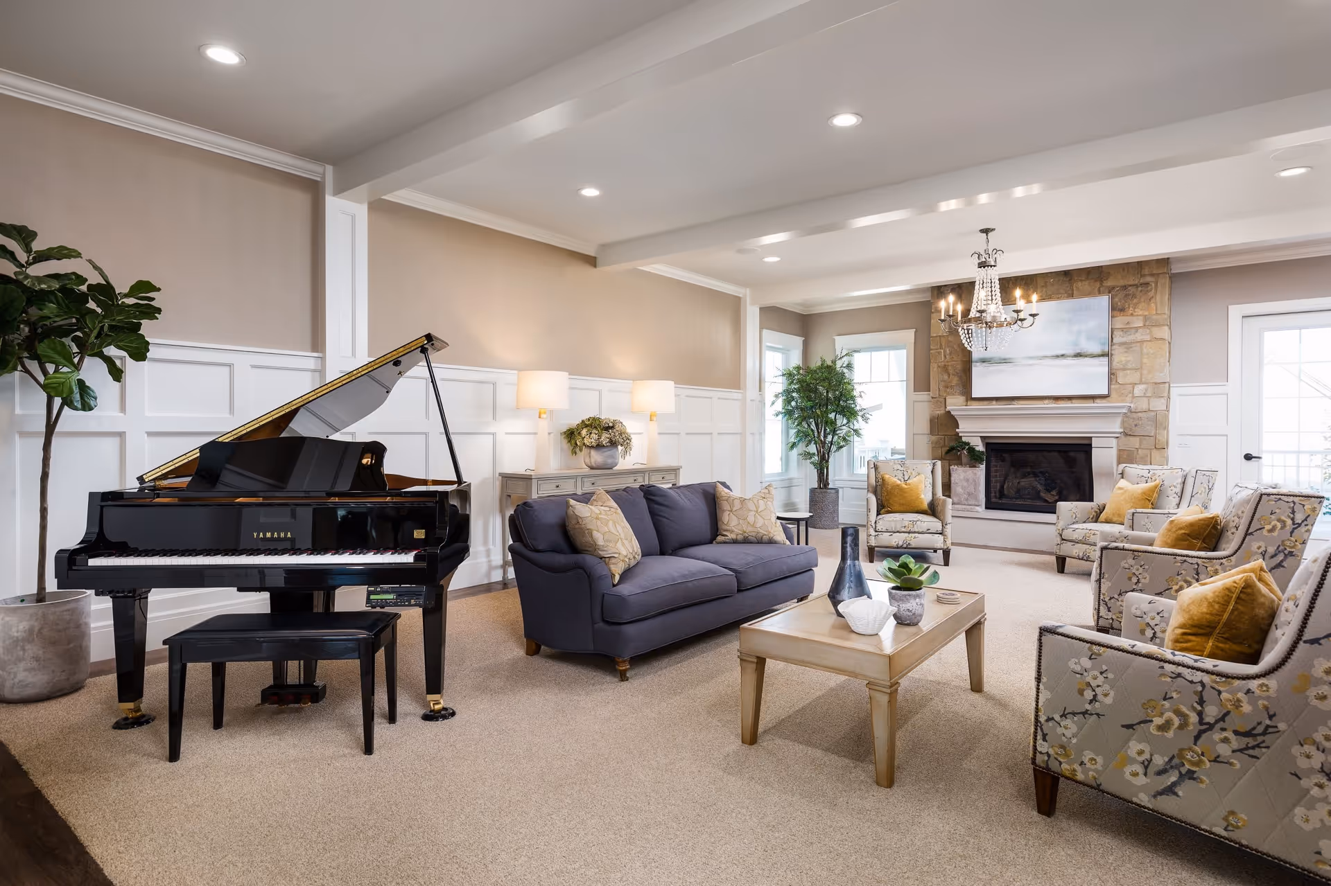 Bright, furnished living room with a grand piano, navy sofa, patterned armchairs, coffee table, and a stone fireplace.