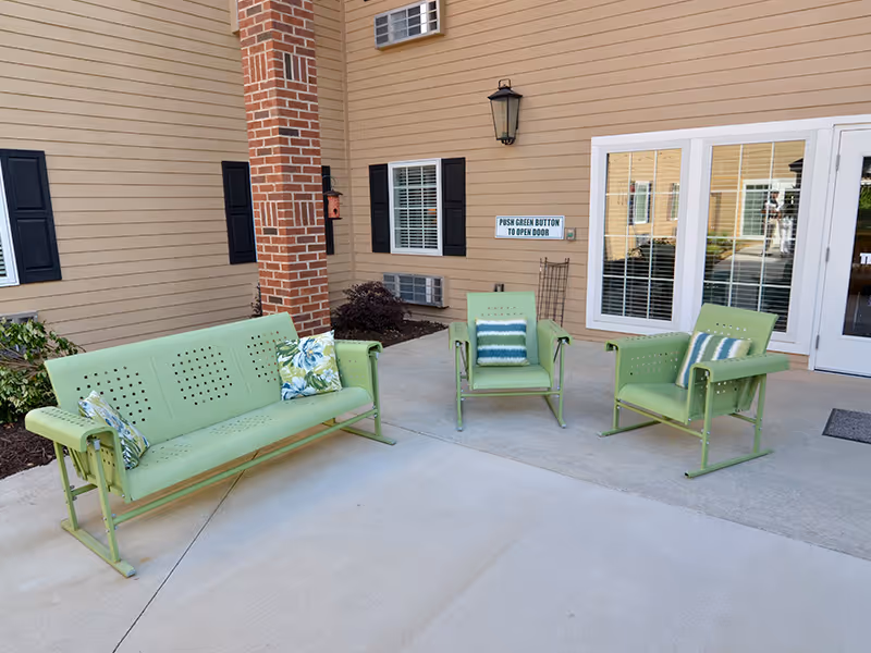 Outdoor patio area with light green metal bench and two matching chairs, each with decorative pillows, situated on a concrete surface near a building with beige siding, black shutters, and a brick column. A sign on the wall instructs to push a green button to open the door.