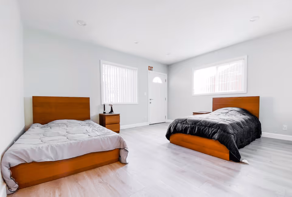A bright, minimalist bedroom with two wooden beds, one with a gray comforter and the other with a black comforter. There are two windows with white blinds, a white door with an exit sign above it, and a small wooden nightstand with a lamp between the beds. The floor is light-colored wood, and the walls are painted white.