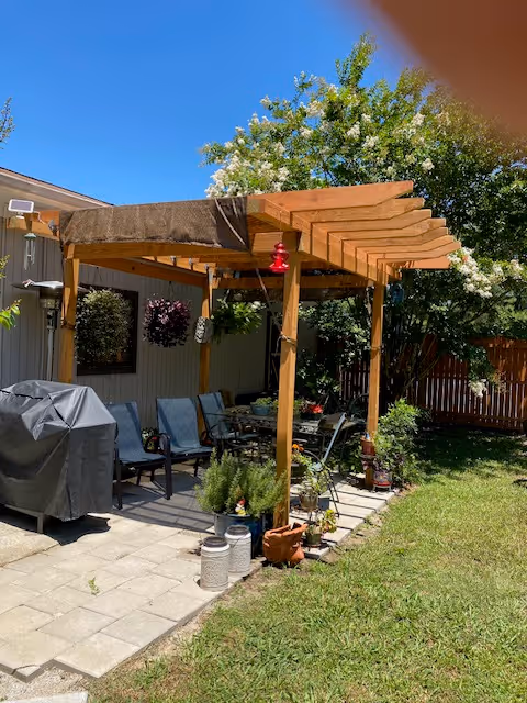 Outdoor patio area with a wooden pergola providing partial shade over a seating area with chairs and a table. There is a covered grill on the left side and various potted plants around the patio. The background shows a wooden fence, green grass, and trees with white flowers under a clear blue sky.