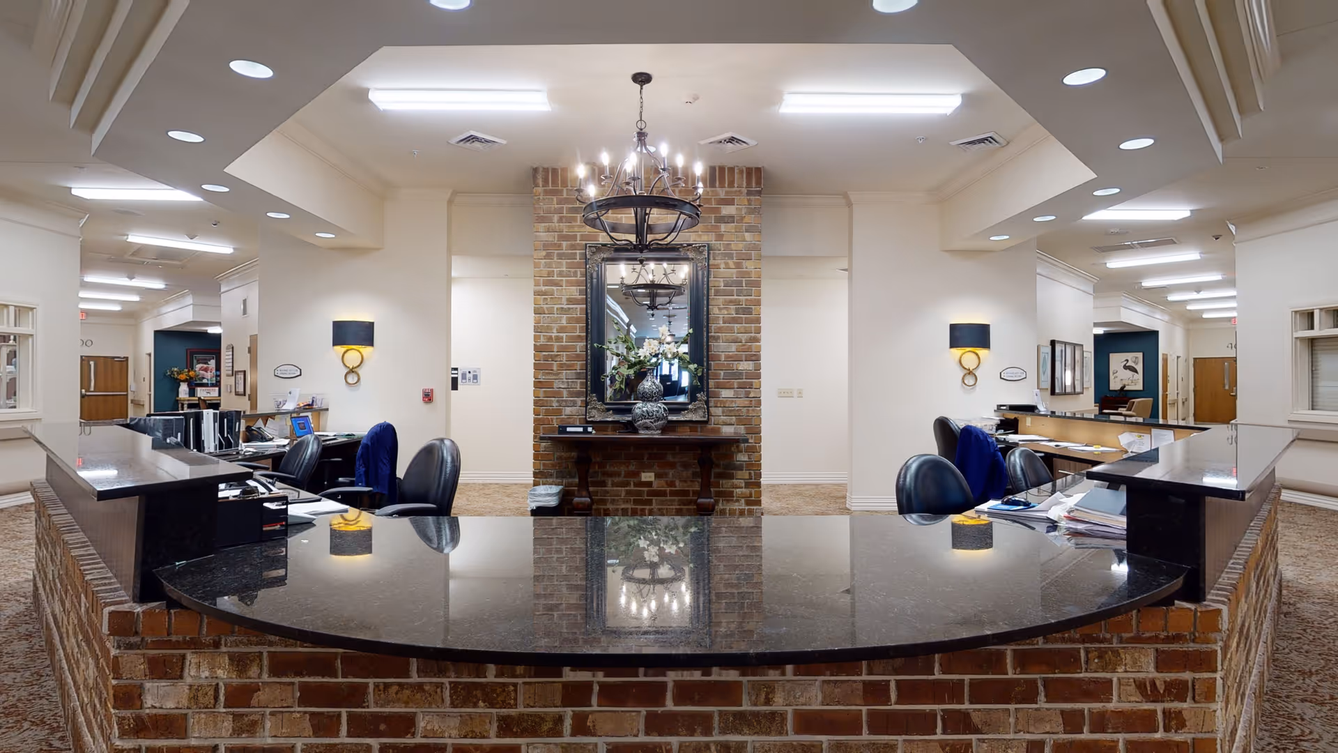Reception area inside Landmark Nursing Center Hammond featuring a curved black granite countertop with brick base, office chairs, desks with paperwork and computers, a brick wall with a large mirror and floral arrangement, and ceiling lights including a chandelier.