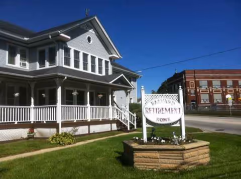 Front exterior of a retirement home with a wraparound porch, sign, and well-kept lawn.