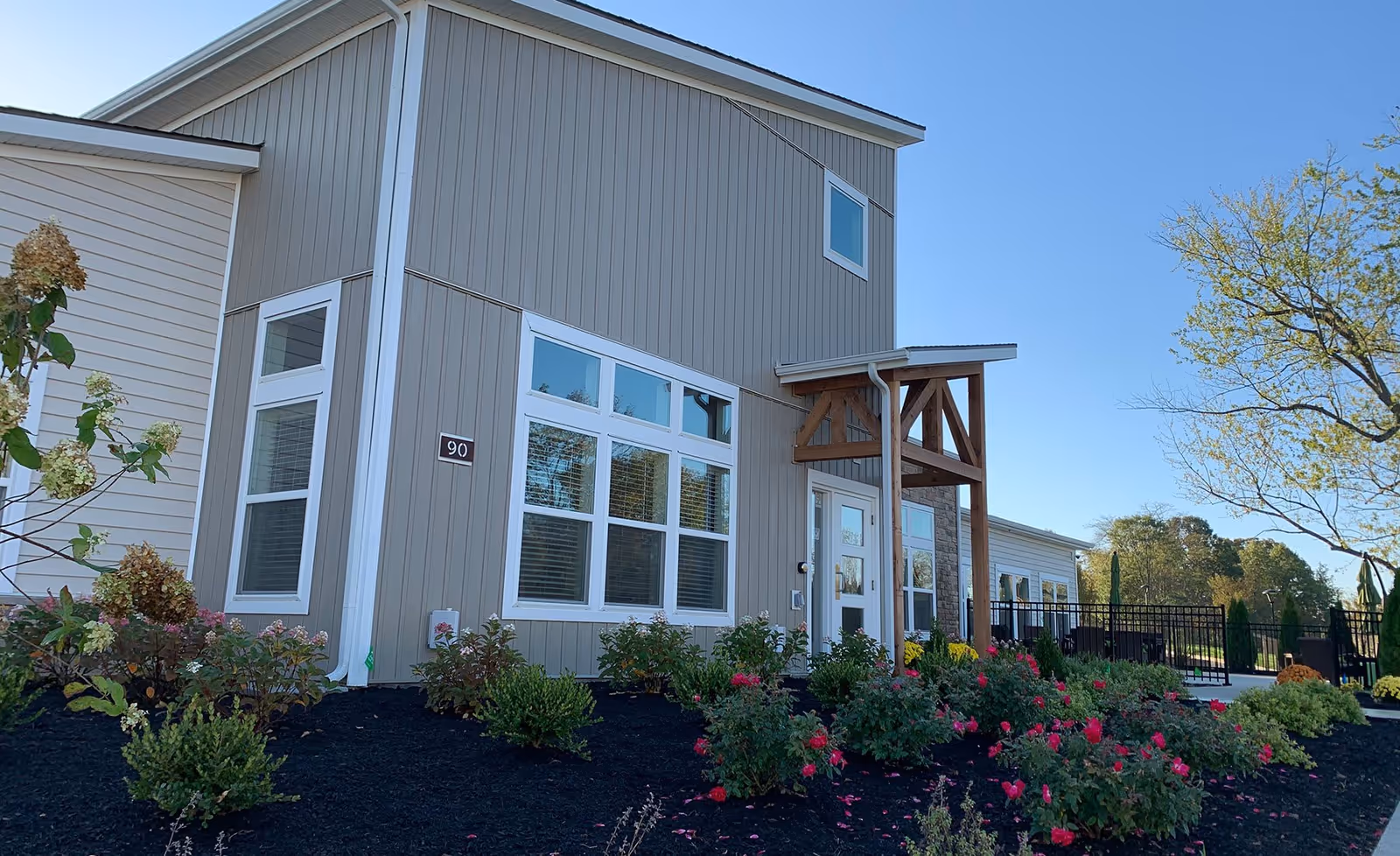 Front exterior of a modern senior living building with large windows, a wooden entry awning, and landscaped flower beds under a clear sky.