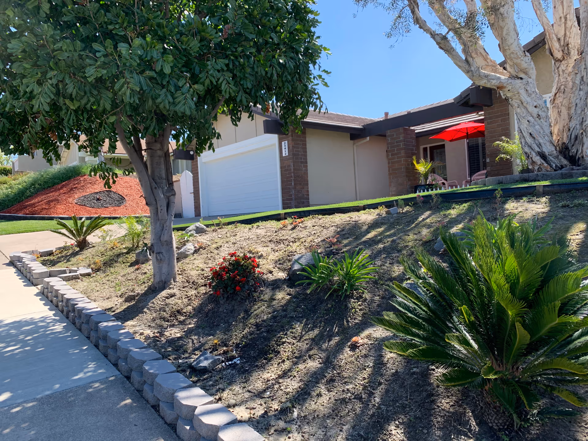 Exterior view of a residential building with a sloped garden area in front. The garden has various plants, including a tree, shrubs, and small flowering plants. There is a concrete sidewalk bordered by a low retaining wall made of stone blocks. The building has a garage with a white door and a covered patio area with a red umbrella and outdoor seating. The sky is clear and blue.
