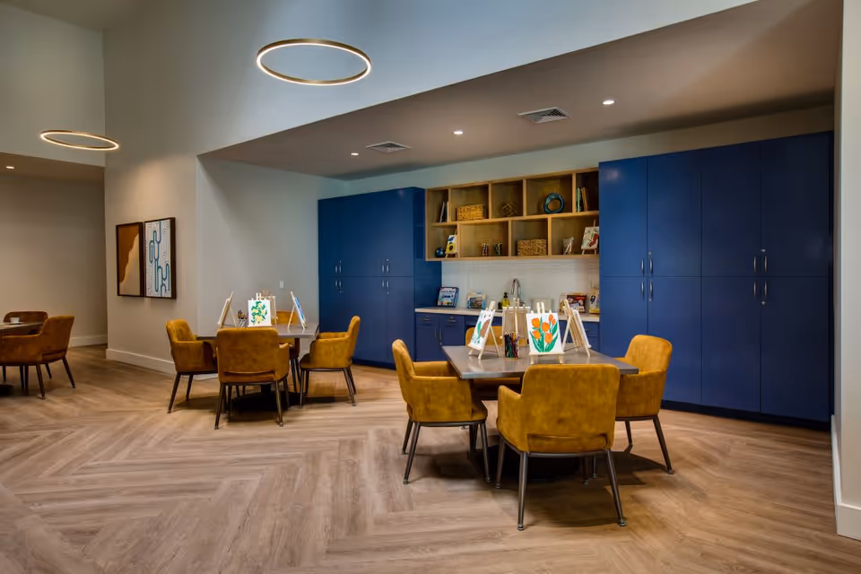 Well-lit communal activity room with tables and mustard-colored chairs arranged around a central table in front of blue built-in cabinets and shelving with art supplies.