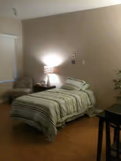 A small bedroom with a single bed covered in striped bedding, a bedside table with a lamp emitting soft light, a cushioned chair in the corner, and a small table with a plant on the right side.