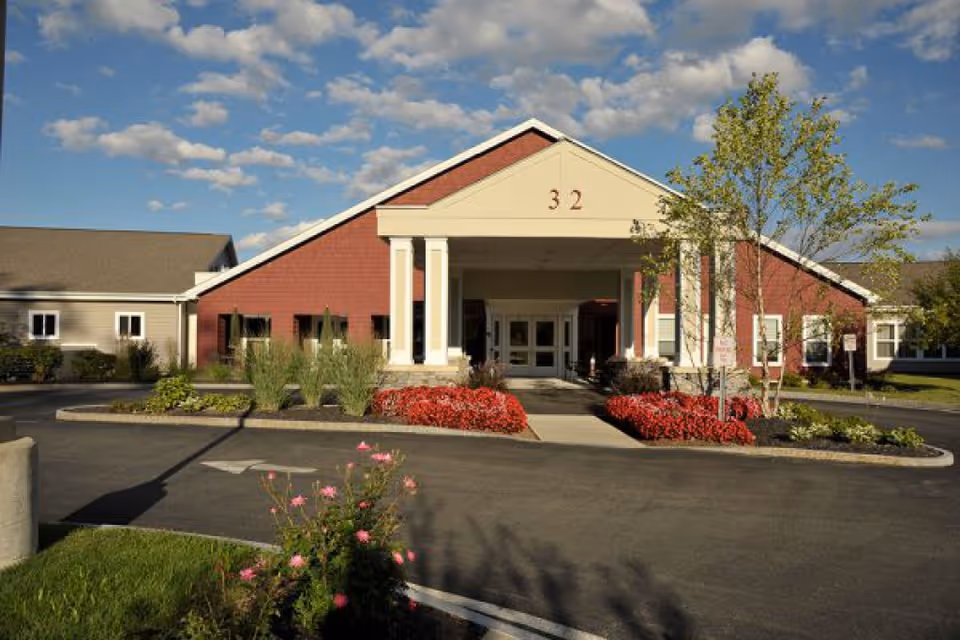 Front exterior view of a single-story building with a red brick facade and beige siding, featuring a covered entrance supported by white columns. The number 32 is displayed on the front gable. There are landscaped flower beds with red flowers and green shrubs along the driveway, and a partly cloudy blue sky overhead.