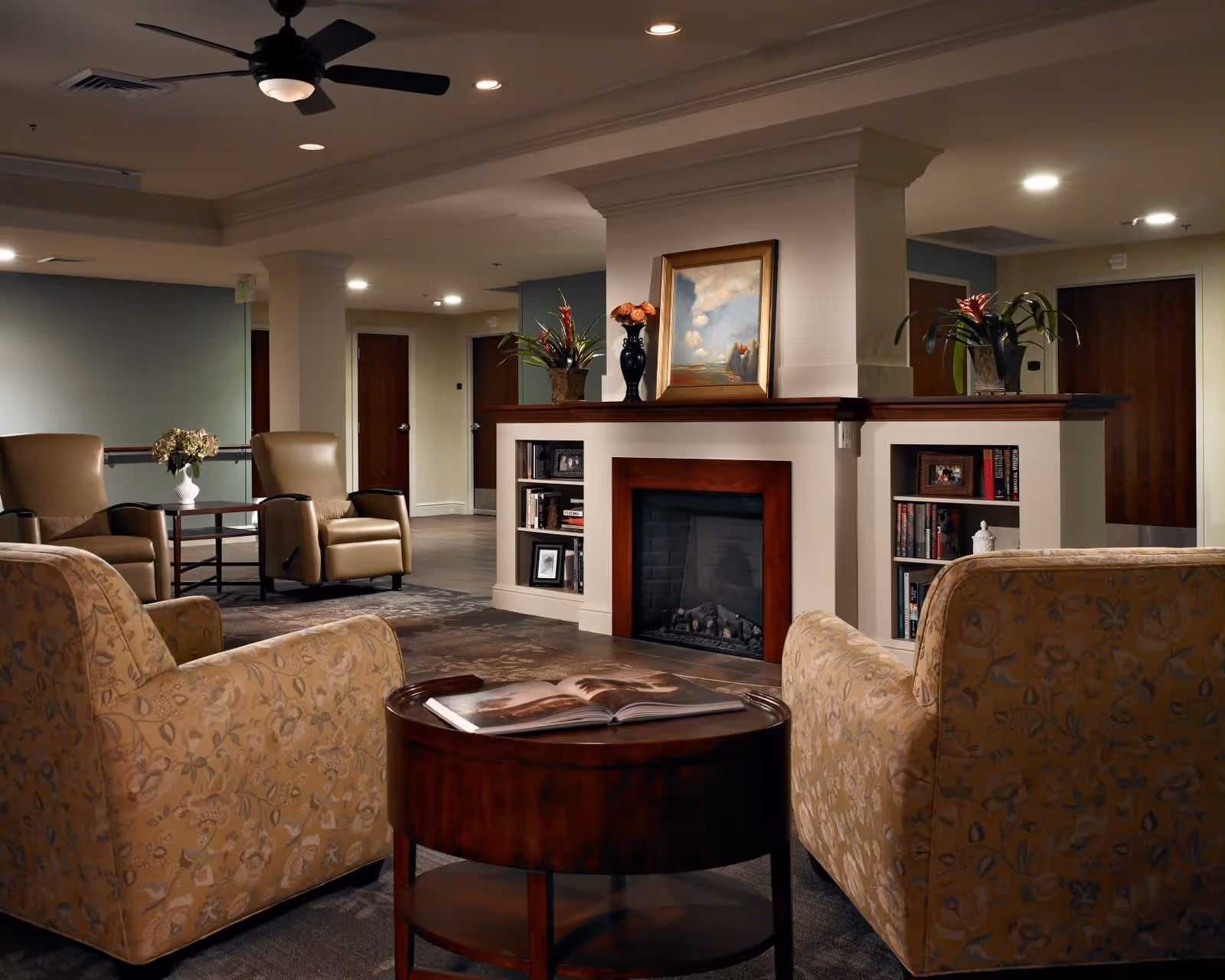 A cozy living room area in a senior living facility featuring a central fireplace with built-in bookshelves on either side. The room has comfortable armchairs and a small round wooden table with an open book on it. There are decorative plants and a framed painting above the fireplace. The ceiling has recessed lighting and a ceiling fan, and there are wooden doors in the background.