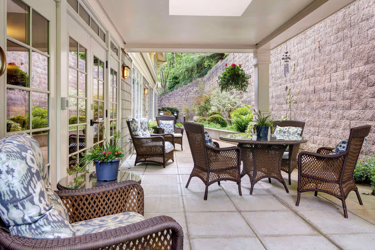 Covered outdoor patio with wicker chairs and a round table beside glass doors and a landscaped retaining wall.