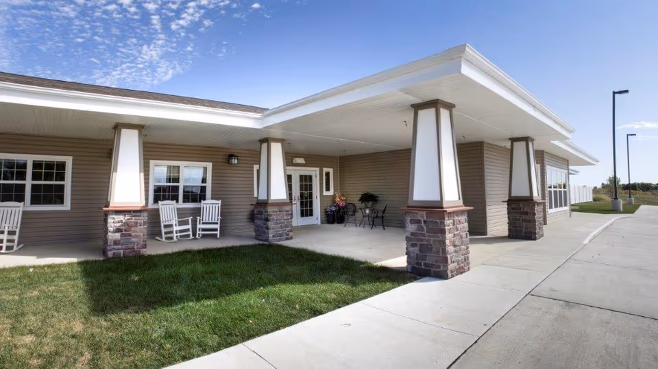 Covered front entrance of a single-story assisted living building with stone pillars, rocking chairs, a concrete walkway and a small lawn under a clear blue sky.