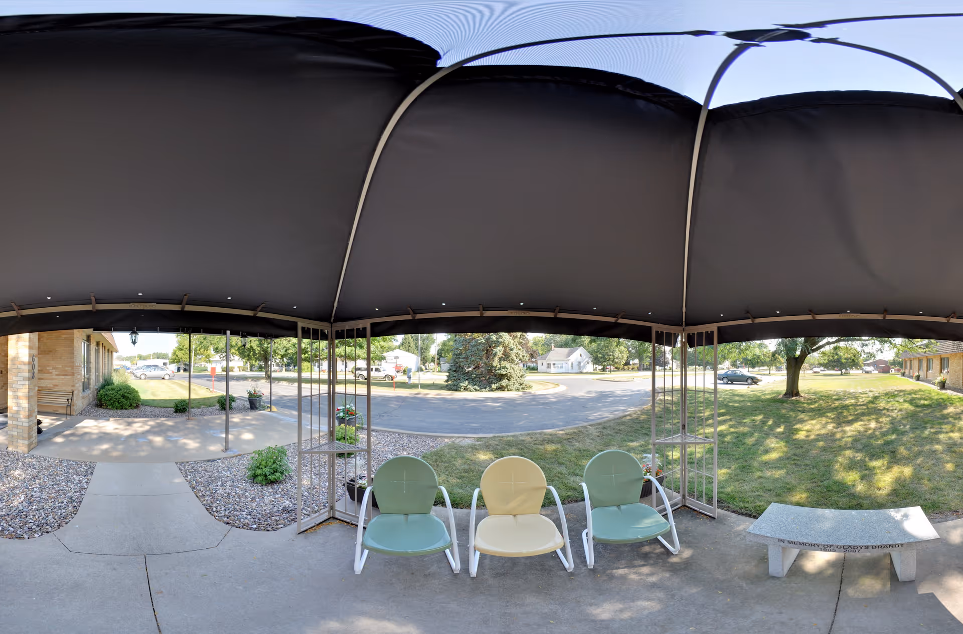 Covered entrance patio with three metal lawn chairs and a stone bench facing a driveway and grassy lawn.