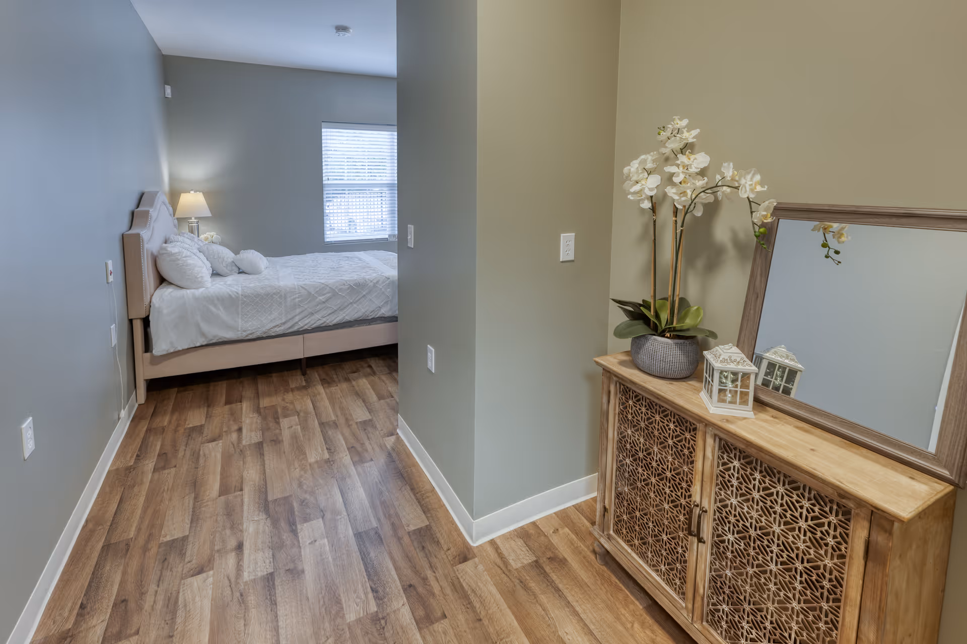 A small hallway opens into a bedroom with a bed on the left and a wooden cabinet topped with a mirror and orchid on the right.