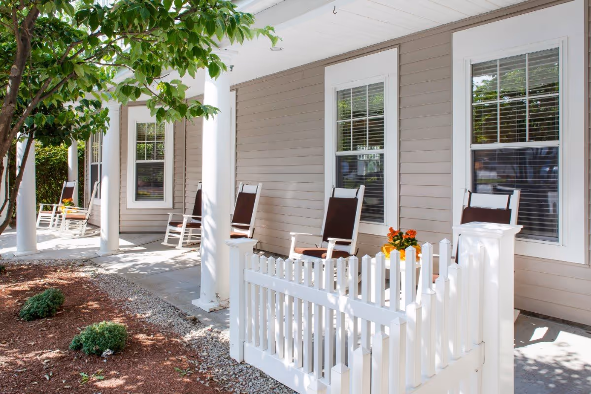 Covered front porch with white columns, rocking chairs, a small white picket fence and potted flowers.